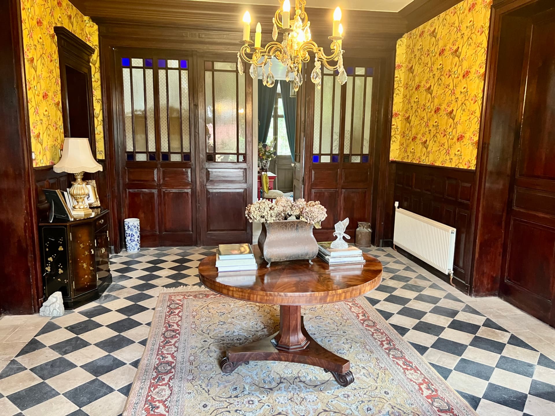 The Grand Entrance Hall with yellow floral wallpaper, wood panelling, chandelier and chequered stone floor